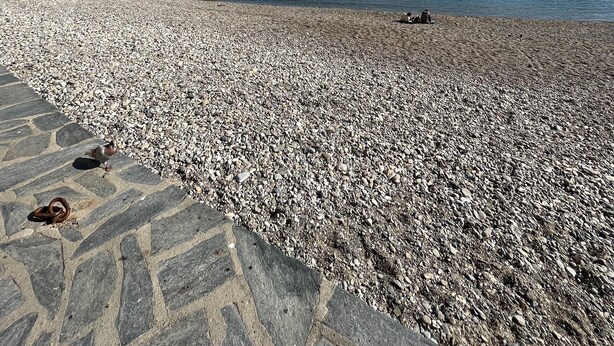 Ein Kiesstrand mit einem historischen Steinbau und blauem, klarem Himmel.