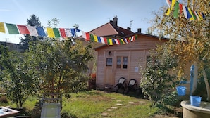 Eine Holzhütte mit einem Ziegeldach, umgeben von einem Garten mit einem gepflasterten Weg, verziert mit bunten Gebetsfahnen.