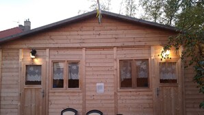 Eine Holzhütte mit drei schwarzen Plastikstühlen davor, zwei Fenstern mit Spitzenvorhängen und einem Schild, das einen privaten Raum anzeigt.