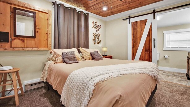 A bedroom with a bed, wooden ceiling, a mirror, a door, and a small stool.