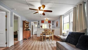 A spacious living area with a wooden floor, a dining table with chairs, a ceiling fan, and a sofa with cushions.