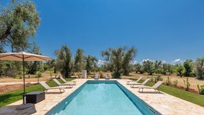 A rectangular outdoor swimming pool with lounge chairs and an umbrella, surrounded by greenery and trees.