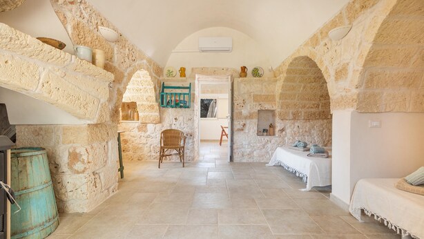 A stone-walled room with a high ceiling, a wicker chair, a bench with towels, and a rustic barrel.