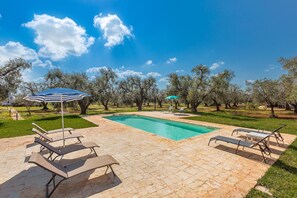 Una piscina con sillones de descanso y sombrillas, rodeada de árboles y un cielo despejado.