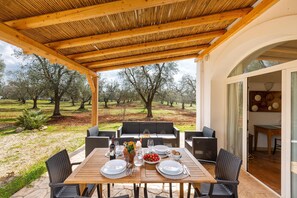 An outdoor dining area with a wooden table set for two, surrounded by wicker chairs, overlooking a grassy landscape with trees.