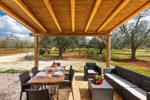 Un patio exterior con una pérgola de madera, suelo de piedra, mobiliario de mimbre y vistas a un olivar.