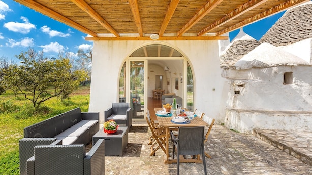 An outdoor patio with a wooden pergola, a dining table set for two, and a white building with a stone roof in the background. - Trullo Palma by Perle di Puglia (San Michele Salentino)