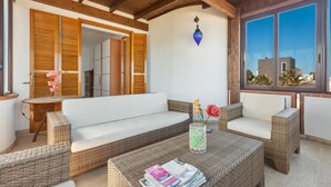 Una habitación con muebles de mimbre, un sofá blanco, una mesa de centro con revistas y un ventanal con vista a edificios.
