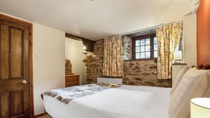 Ein Schlafzimmer mit einer Steinswand, einer Holztür, einem Bett mit weißer Bettdecke, einem Schminktisch, einem Spiegel und einem Fenster mit blumigen Vorhängen.