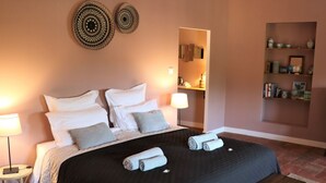 Una cama bien hecha con ropa de cama blanca y gris, dos adornos en la pared y una mesita de noche de madera con una lámpara.