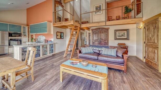 Amplia sala de estar con un entrepiso con cama, muebles de madera y una cocina al fondo.