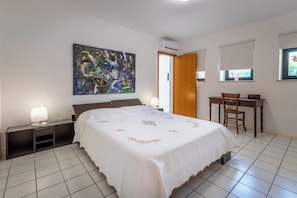 A bedroom with a bed, a desk, a chair, a nightstand, a lamp, and a painting on the wall.