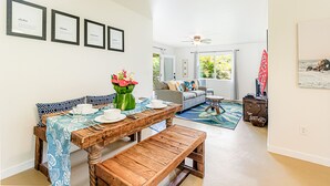A dining area with a wooden table and benches, a sofa, and a coffee table. There are framed pictures on the wall and a surfboard leaning against the wall. - Jungle Bungalow - A lovely space to stay for island fun! (Keaau)