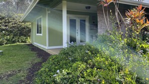 A single-story house with a green exterior, a front porch, and a small garden area with various plants and flowers. - Jungle Bungalow - A lovely space to stay for island fun! (Keaau)