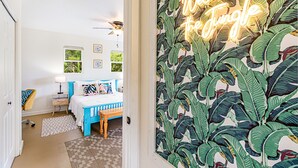 A bedroom with a bed, bedside tables, a ceiling fan, and a wall with a tropical leaf pattern and a neon sign. - Jungle Bungalow - A lovely space to stay for island fun! (Keaau)