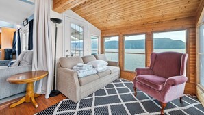 Ein gemütliches Zimmer mit Holzbalkendecke, einer Couch, einem Sessel und großen Fenstern mit Blick auf ein Gewässer.