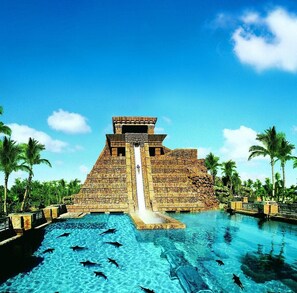 A large pyramid structure with a water slide leading to a pool surrounded by palm trees.