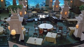 Un área de comedor interior con mesas y sillas, ventanas amplias y pilares decorativos.