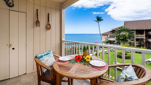 Une table en bois, sur laquelle sont posés une assiette blanche et une fleur rouge, est placée sur un balcon d’où l’on aperçoit l’océan et, au loin, un bâtiment.