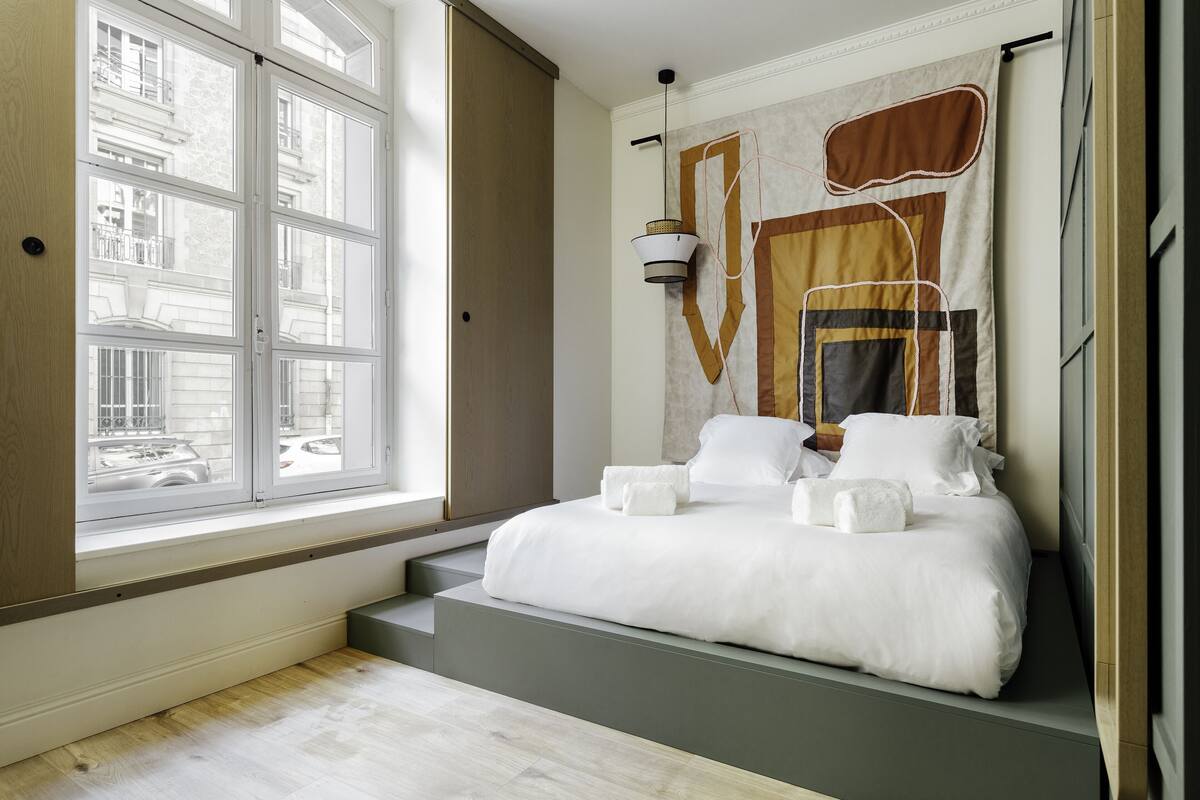 A bedroom with a large window, a bed with white linens, and a unique wall tapestry.