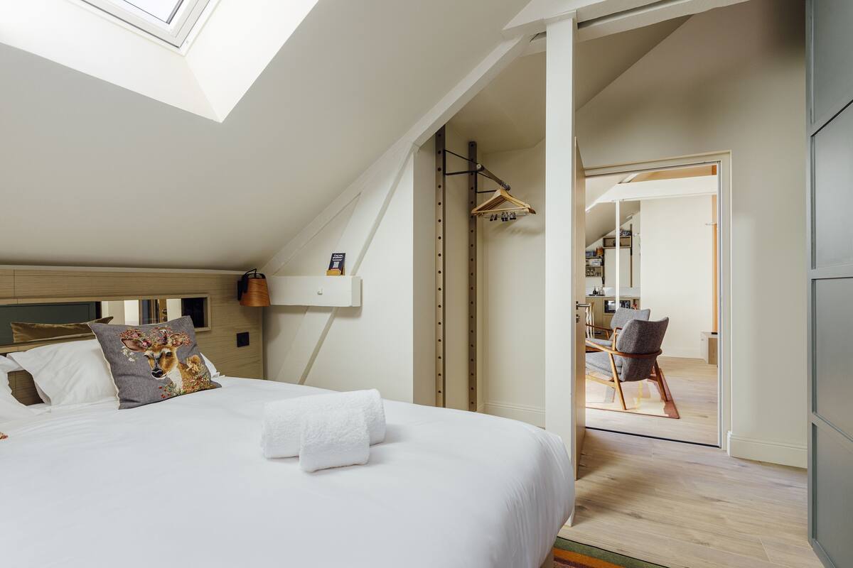 A bedroom with a skylight, a bed with white linens, a wooden headboard, and a view into the adjacent room.
