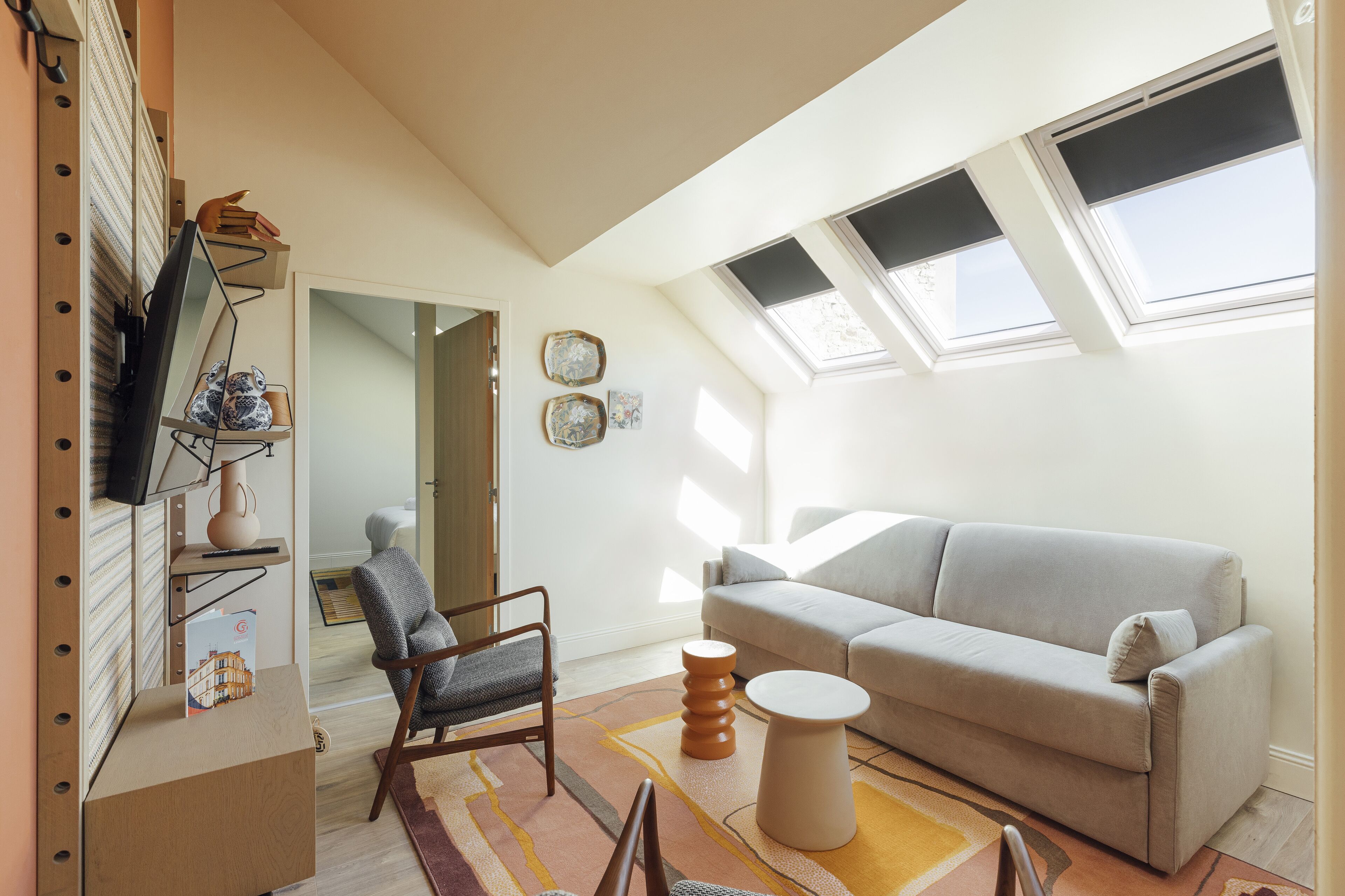 Una sala de estar moderna con un sofá, un sillón y una mesita redonda. Hay un televisor sobre un mueble bajo, una estantería y una ventana con persianas.