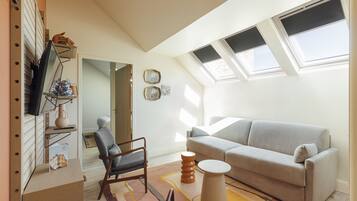 Una sala de estar moderna con un sofá, un sillón y una mesita redonda. Hay un televisor sobre un mueble bajo, una estantería y una ventana con persianas.