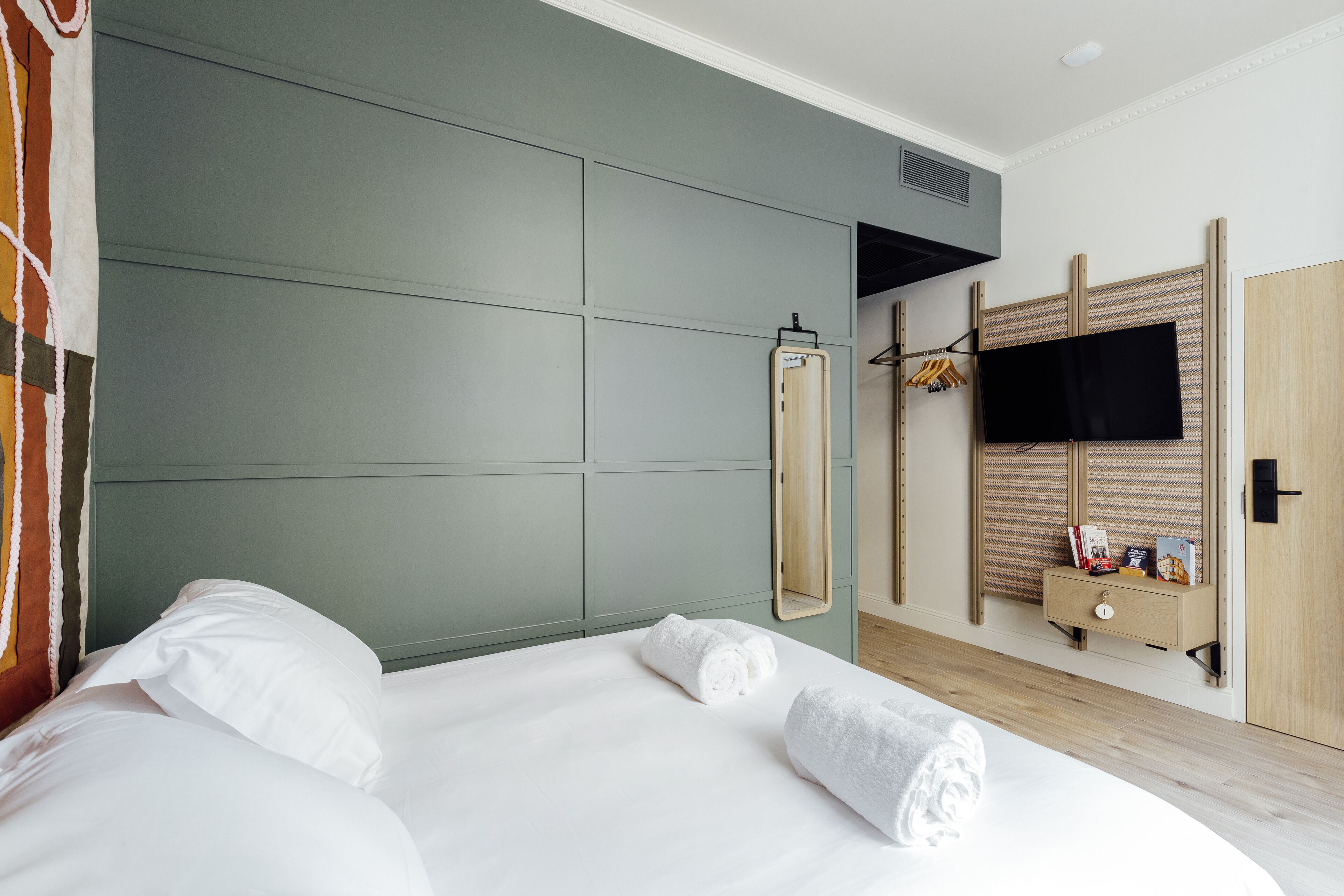 Une chambre moderne avec un grand lit, une armoire en bois, un téléviseur fixé au mur et une petite table de chevet avec des livres et une lampe.