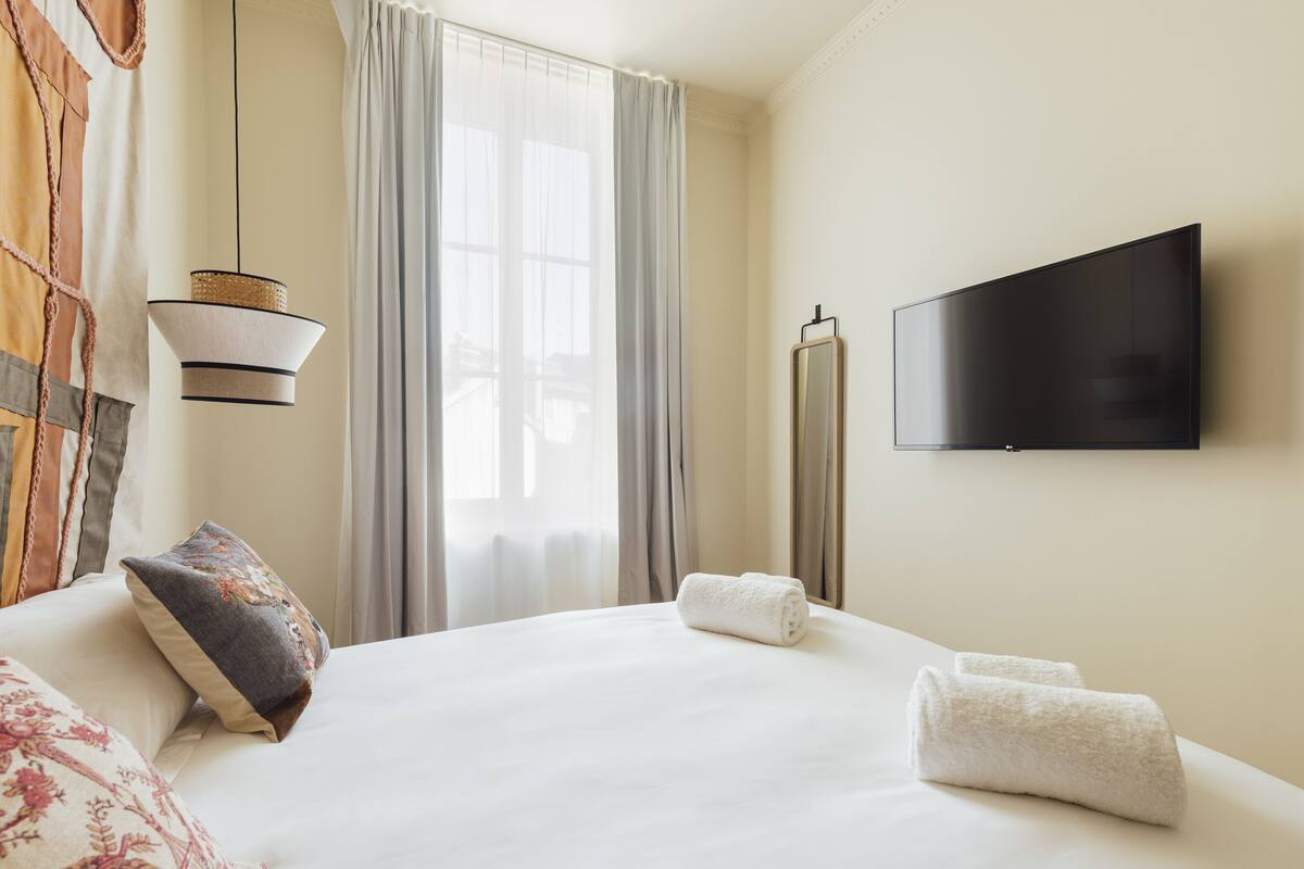 A hotel room with a large bed, a flat-screen TV mounted on the wall, a window with curtains, and a hanging light fixture.