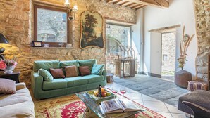 A cozy living room with a stone wall, a green sofa, a coffee table with books and fruit, and a large window. - Podere Le Corone 14, Emma Villas (Perugia)