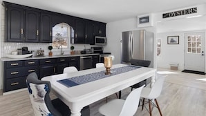 Une cuisine moderne avec des meubles de couleur sombre, un plan de travail blanc, des appareils en acier inoxydable et un coin repas avec une table et des chaises blanches.