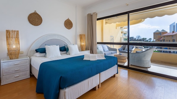 Quarto de hotel moderno com uma cama grande, uma mesa de cabeceira, uma cadeira e vista para a cidade através de amplas janelas.