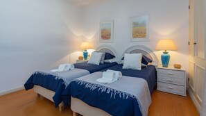 Uma cama de casal com colcha azul e almofadas brancas, duas mesas de cabeceira com abajures e um pequeno vaso decorativo.