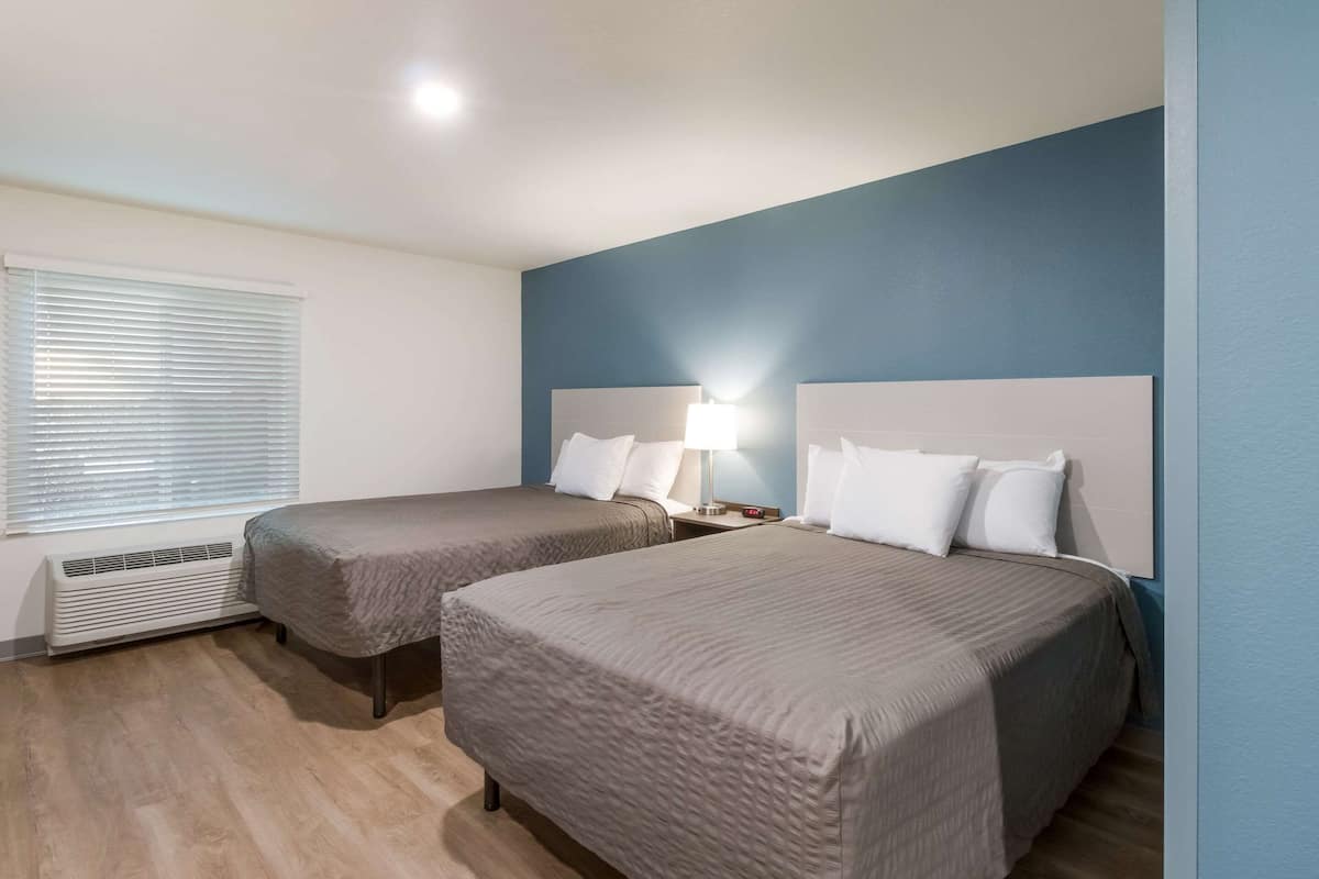 A hotel room with two beds, a window with blinds, a bedside lamp, and a wall heater.