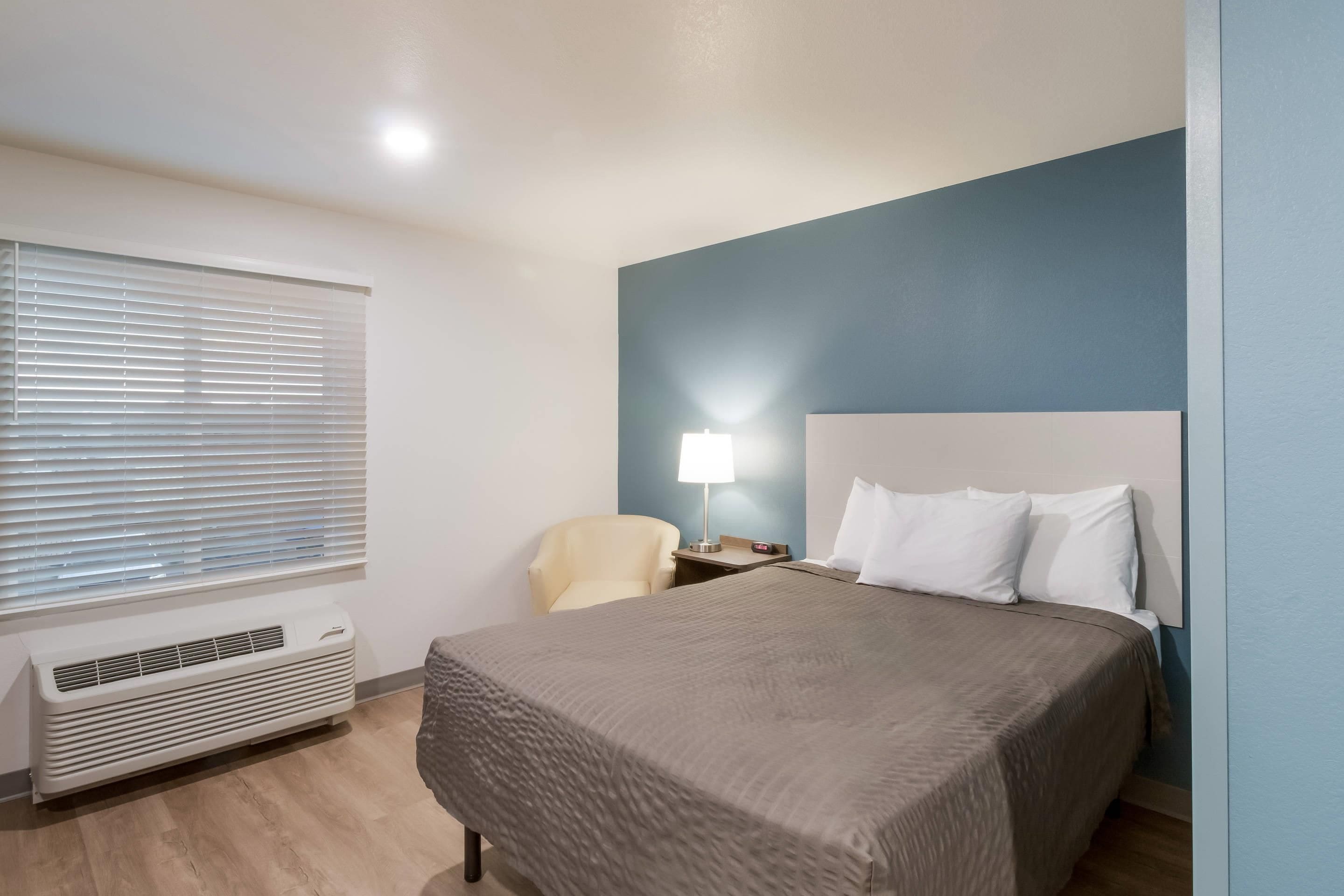 Una habitación de hotel con cama, una silla, mesita de noche con lámpara y ventana con persianas.
