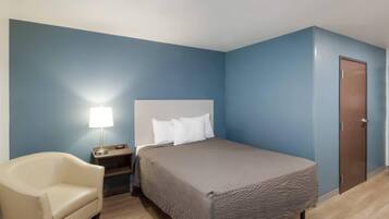Una habitación de hotel con una cama, una silla, una mesita de noche con una lámpara y una puerta.
