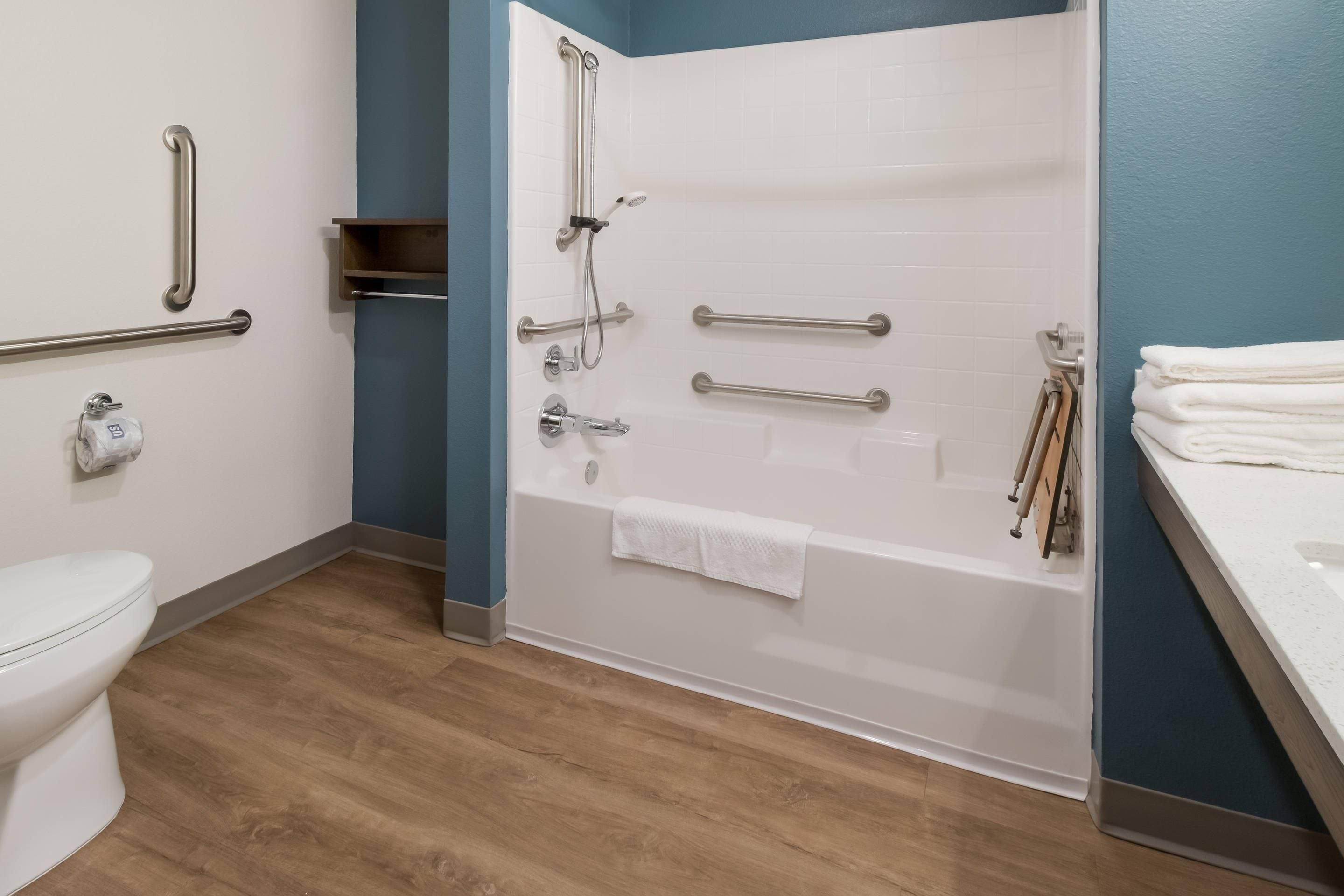 Une salle de bain avec des toilettes blanches, une baignoire avec douche et un lavabo. Des barres d’appui ont été installées pour plus de sécurité.