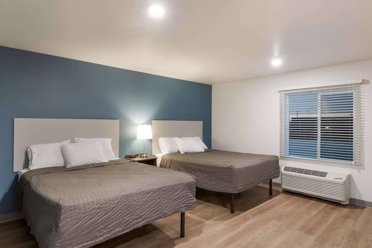 A hotel room with two beds, a nightstand with a lamp, a window with blinds, and a wall-mounted air conditioner.