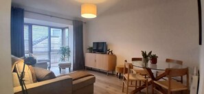 Ein Wohnzimmer mit einer Couch, einem Esstisch mit Stühlen, einem Fernseher auf einer Kommode und einem großen Fenster mit Jalousien.