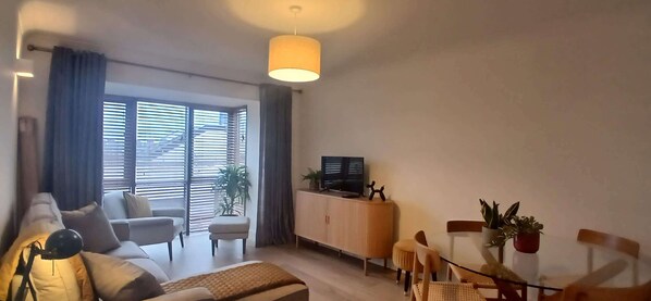 A living room with a sofa, ottoman, and a dining table with chairs. There is a TV on a stand and a plant on the table.