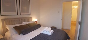A neatly made bed with pillows and a rolled towel, a bedside lamp, and a door leading to another room.