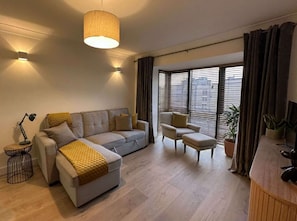 Ein modernes Wohnzimmer mit einer Couch, einem Sessel, einem Hocker, einer Lampe und einem großen Fenster mit Jalousien.