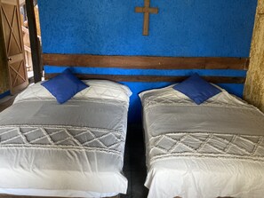 Two single beds with blue pillows sit against a blue wall, adorned with a wooden headboard and a wooden cross.
