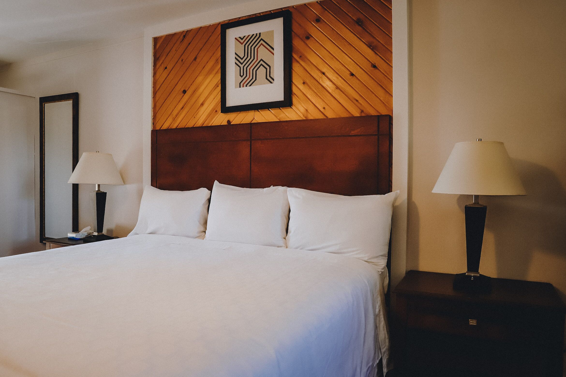 A hotel room with a large bed, two bedside tables with lamps, a wooden headboard, and a framed picture above the bed.