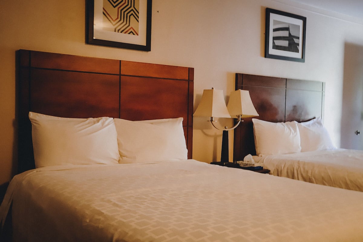 Habitación de hotel con dos camas, cada una con cabecero de madera, ropa de cama blanca y una mesita de noche con lámpara.