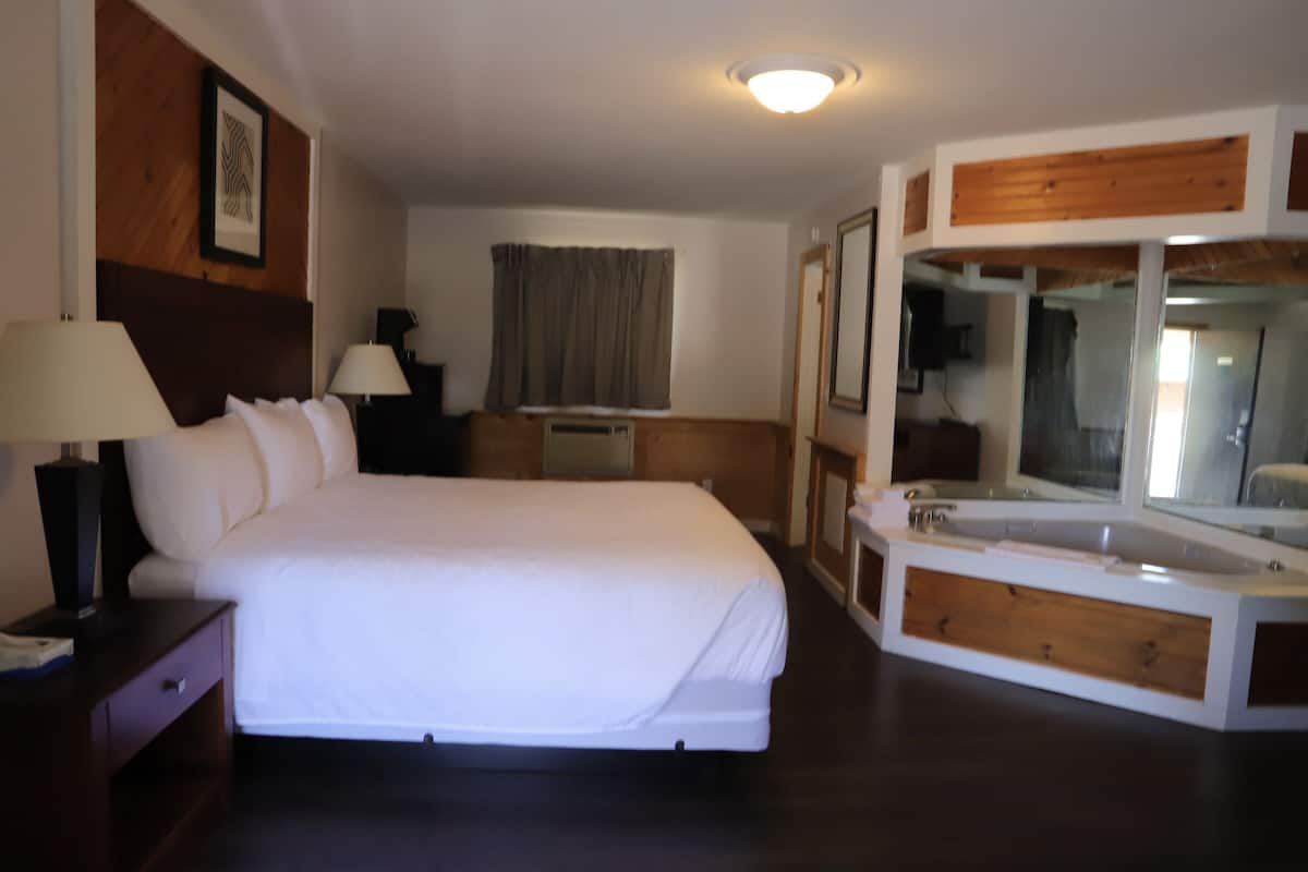 Habitación de hotel con una cama grande, una mesita de noche con lámpara, un espejo y una bañera integrada.