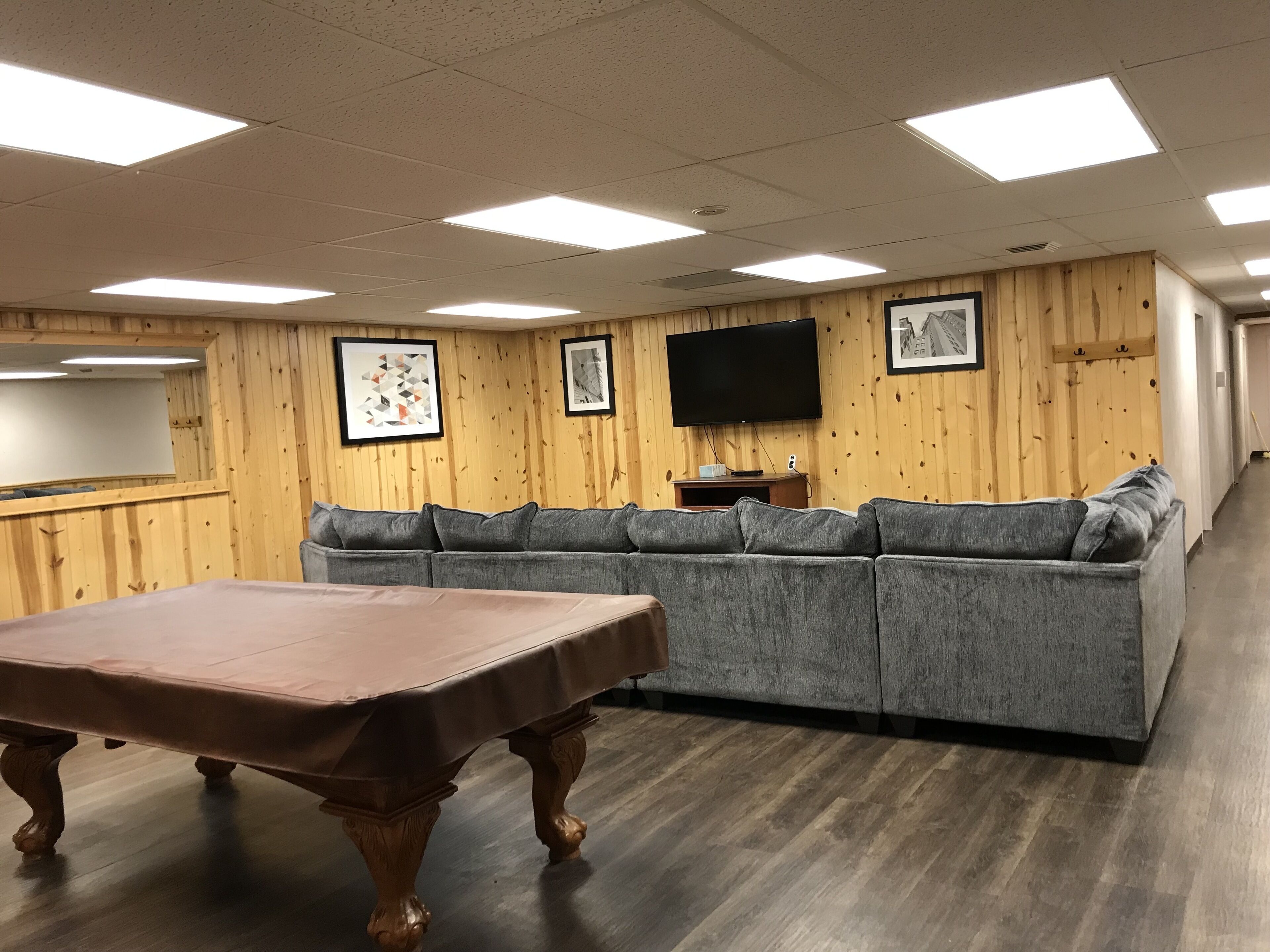 Een kamer met een wand van houten panelen, een grijze bank, een houten tafel en een flatscreen televisie die aan de muur is bevestigd.