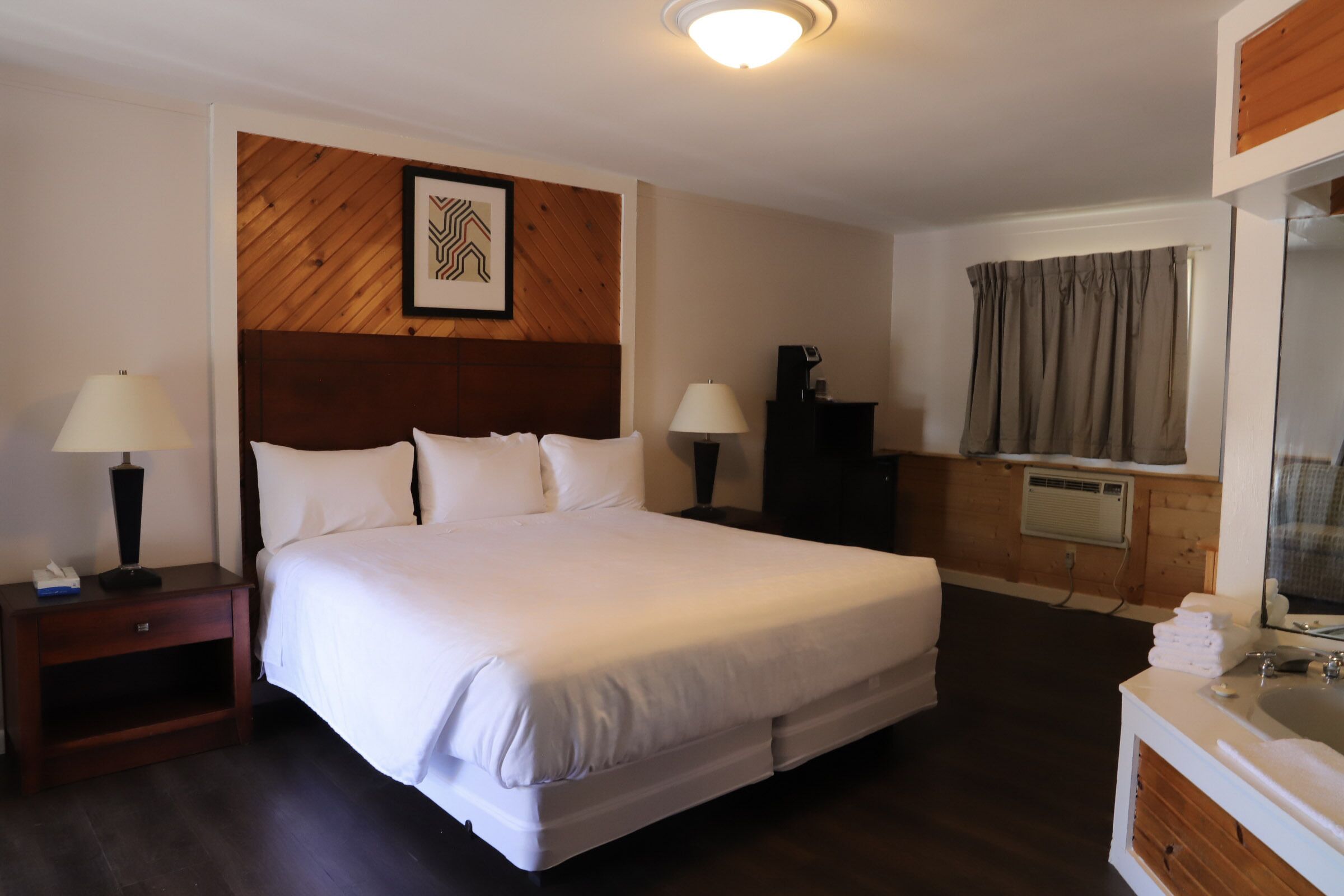 Una cama bien hecha en una habitación de hotel, con cabecera de madera, dos mesitas de noche con lámparas, un mueble bajo y una ventana con cortinas.