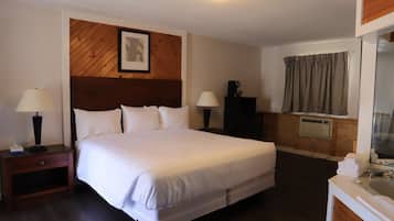 Una cama bien hecha en una habitación de hotel, con cabecera de madera, dos mesitas de noche con lámparas, un mueble bajo y una ventana con cortinas.