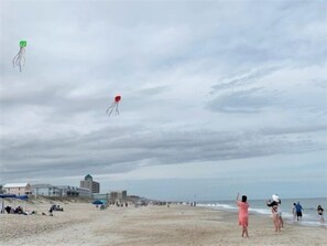 A beach with people flying kites, a sandy shore, and buildings in the background.